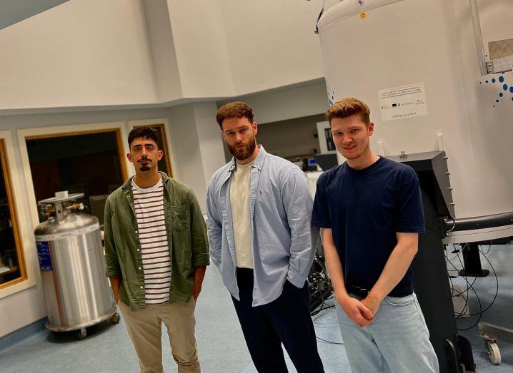 Three young men who co-founded Meta-Flux stand next to each other in an indoor space with a giant cistern behind them.