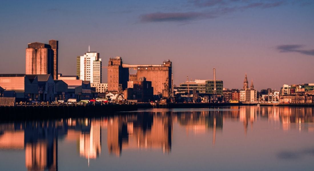 Buildings of Cork city, with reflections of the buildings visible on a river, with a sunset sky in the background.