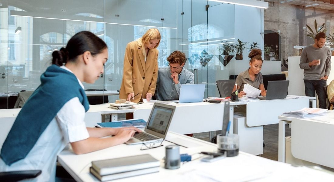 A group of people in an office, some are sitting at computers while others are standing around.