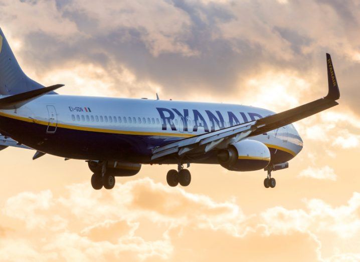 A plane flying in the sky with the Ryanair logo on its side, and the light of the sun visible on clouds in the background.