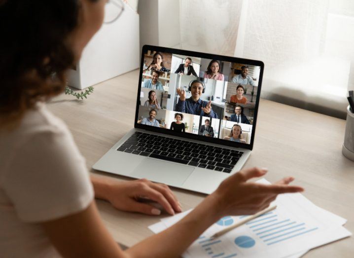 A person speaking to a group of individuals on a laptop screen. The laptop is on a wooden table in a room.
