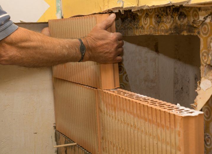 A person putting in a new wall in front of a brick wall, with evidence of damp marks on parts of the older material. Used as a concept for retrofitting works.