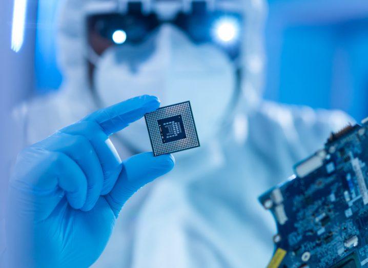 A person in a white lab coat and a mask holding a small semiconductor in a blue gloved hand..