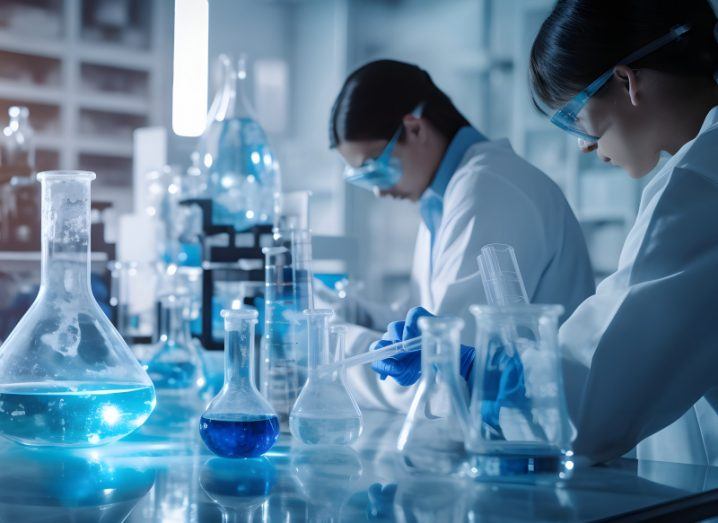 Two people in white lab coats in a medical room, working at a table that has various containers for blue liquids.