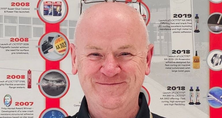 A man smiles while standing in front of an information board. He is Barry Burns, a technical lead at Henkel.