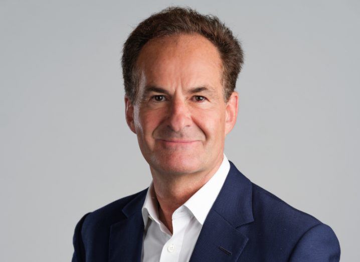 A man in a navy suit, in front of a grey background. He is Brian Menell of TechMet.