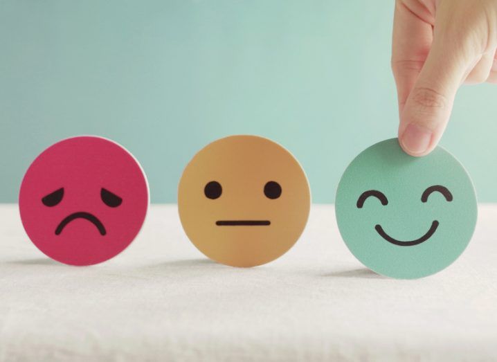 A hand choosing a green smiley face cut-out over an orange straight face and red sad face.