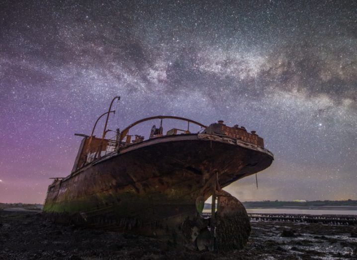 A rusty ship laying under a night sky, which shows large amounts of stars and gases from space.