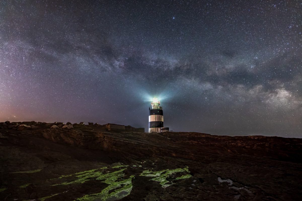 Image of a lighthouse with a light shining from the top and the night sky above it. Stars and gases are visible in the sky above.