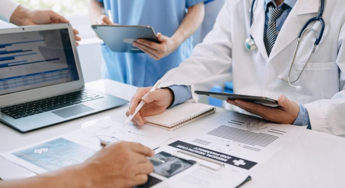 A group of medical staff stand around a desk full of medical records and documents, as well as a laptop.