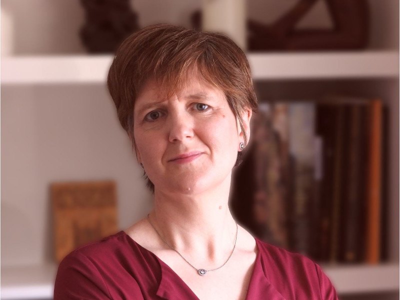 A woman with short hair wearing a dark red top smiles at the camera. In the background is an out-of-focus bookcase.