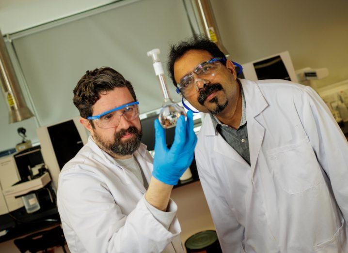 Kris O'Dowd in a lab with his supervisor Prof Suresh Pillai. They are wearing white lab coats and goggles. Kris has blue gloves and is holding up a test tube.