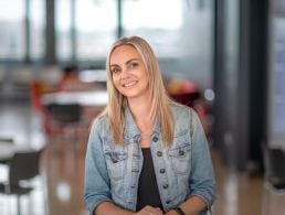 A woman wearing a maroon jumper stands smiling with a wall behind her. On the wall, there are two shelves with toys and plants on them. She is Lynn D'Arcy, a data centre engineer at Yahoo.