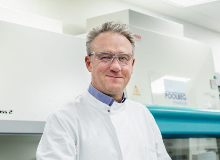 A man in a lab coat and safety glasses smiles at the camera.