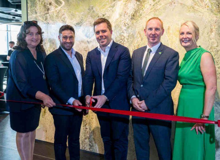 Three men and two women stand in front of a red women smiling at the camera. The man in the centre is holding a scissors to officially launch the new Signify Health centre in Galway.