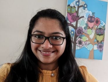 A woman with dark hair wearing glasses and a yellow top smiles at the camera with a wall behind her. There is a painting on the wall depicting owls sitting in a tree.