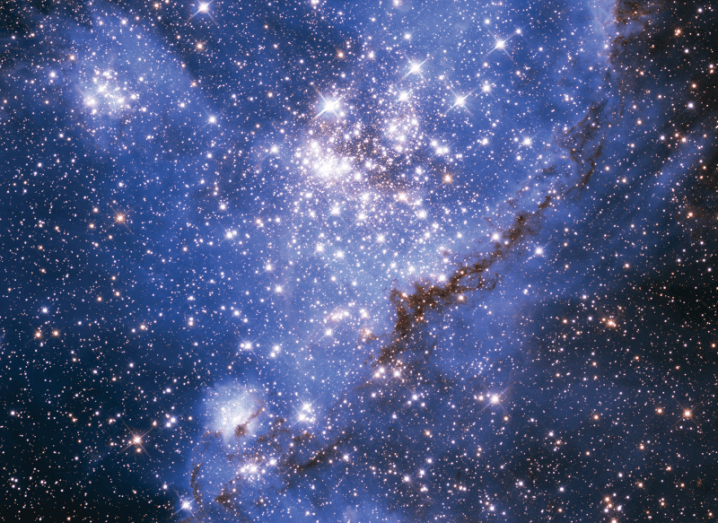 A section of the star-forming nebula NGC 346 taken with the Hubble telescope. The outer areas of the image are dark with bright stars and the centre of the image is purple and blue and grey with brighter dots visible.